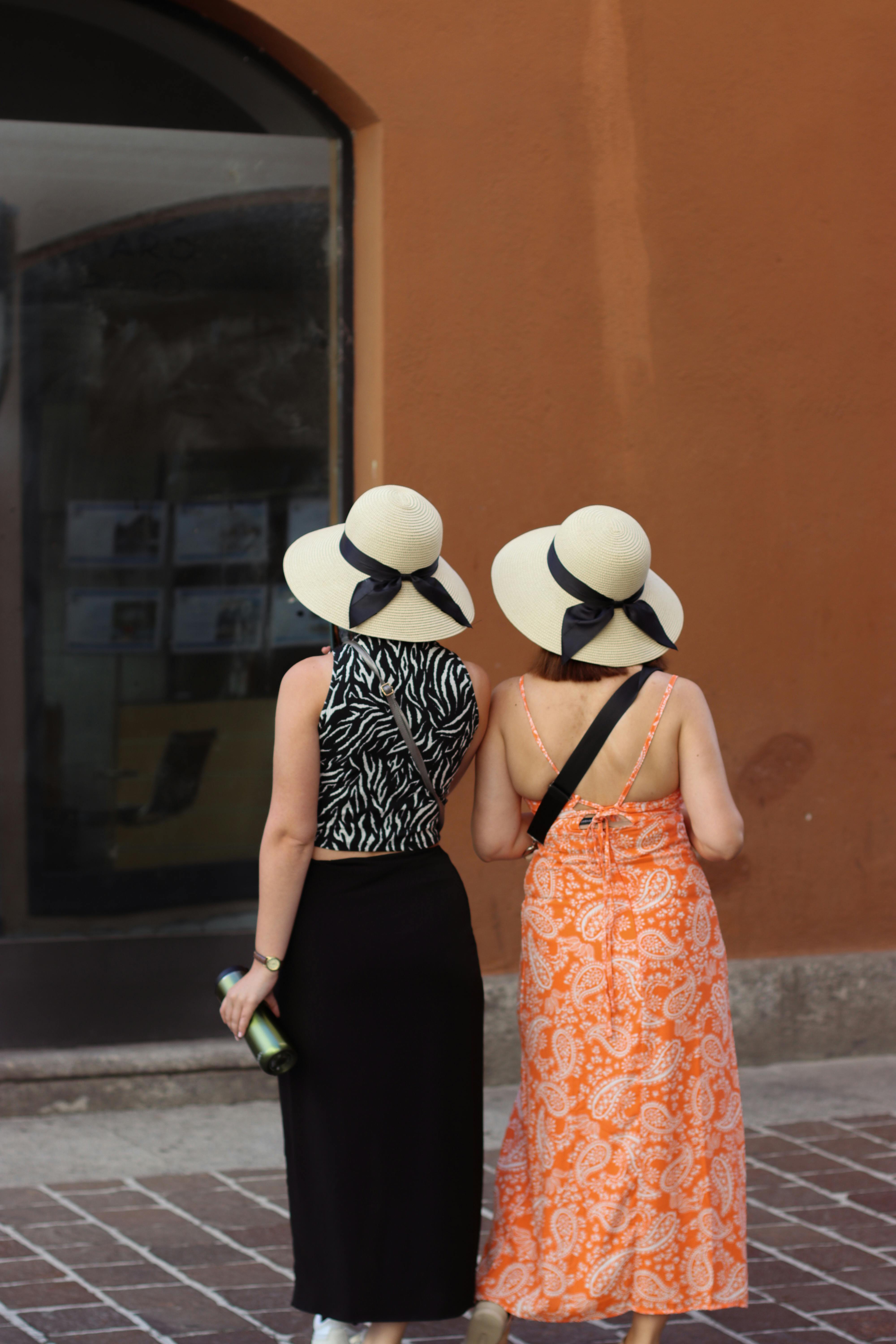Two women in stylish summer dresses and hats walking in Como, capturing Italian street fashion and elegance.