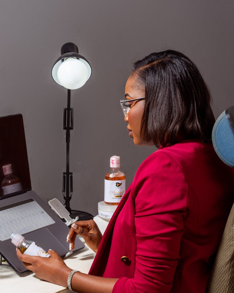 Woman Sitting At A Desk Looking At A Bottle Of Skin Care Cosmetic In Her Hand