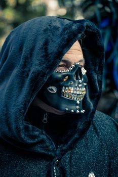 Portrait of a man in a hood wearing a spooky skull mask decorated with spikes and rhinestones, capturing a Halloween vibe.