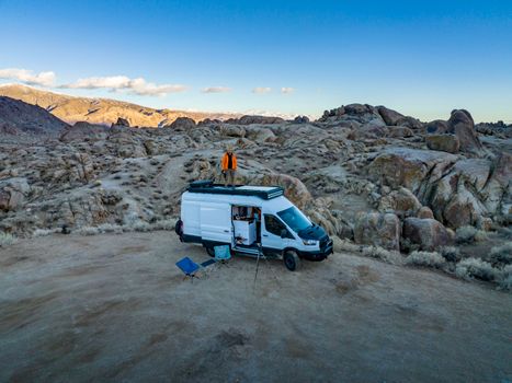 Aerial shot of a man standing on a camper van surrounded by rocky terrain in a remote desert setting.