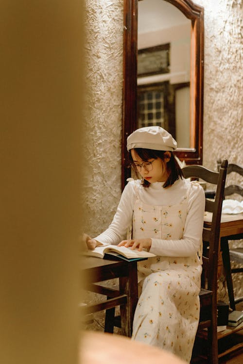 Woman in Hat and White Dress Sitting by Table and Reading