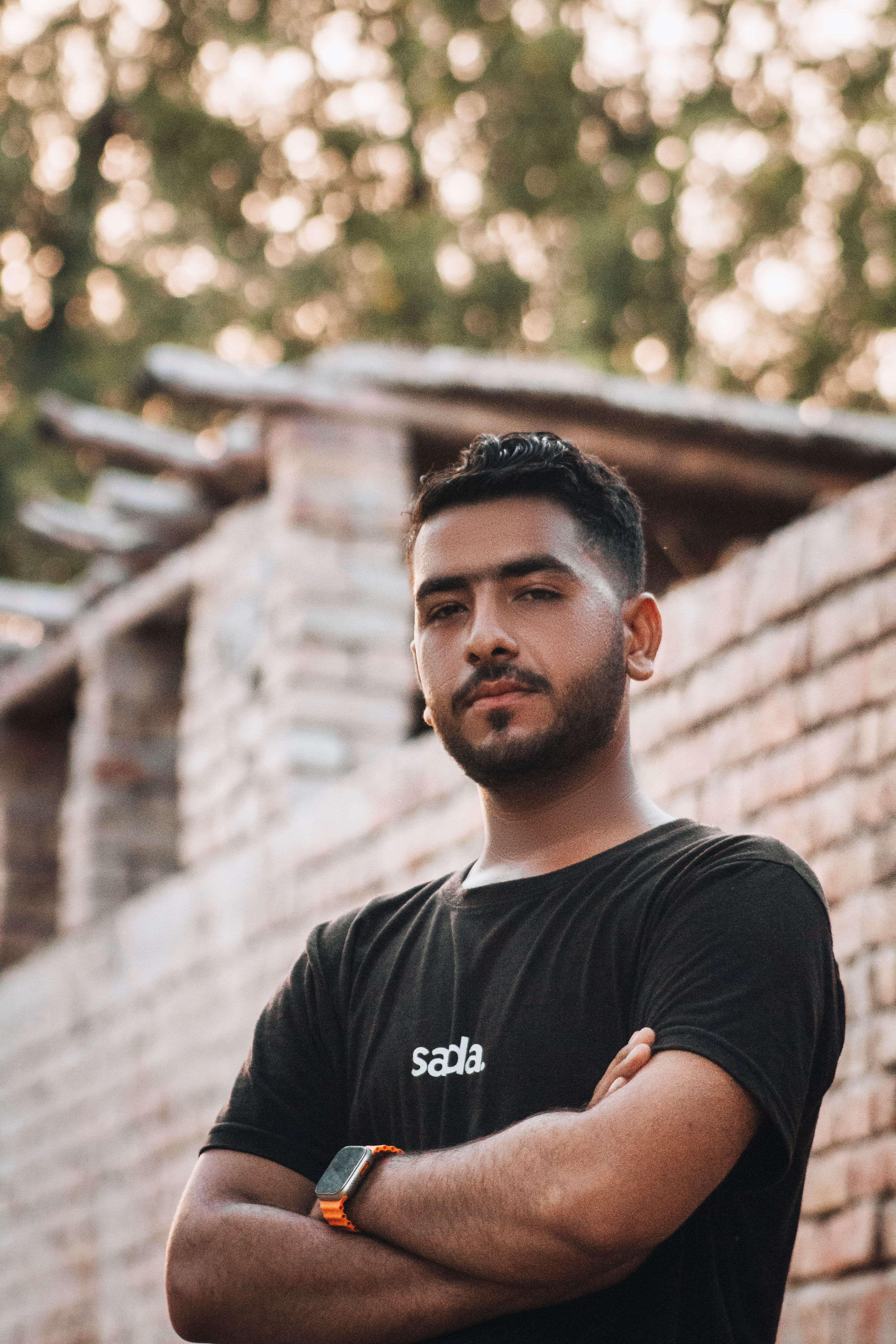 Young man in a black t-shirt poses confidently against a rustic brick wall outdoors.