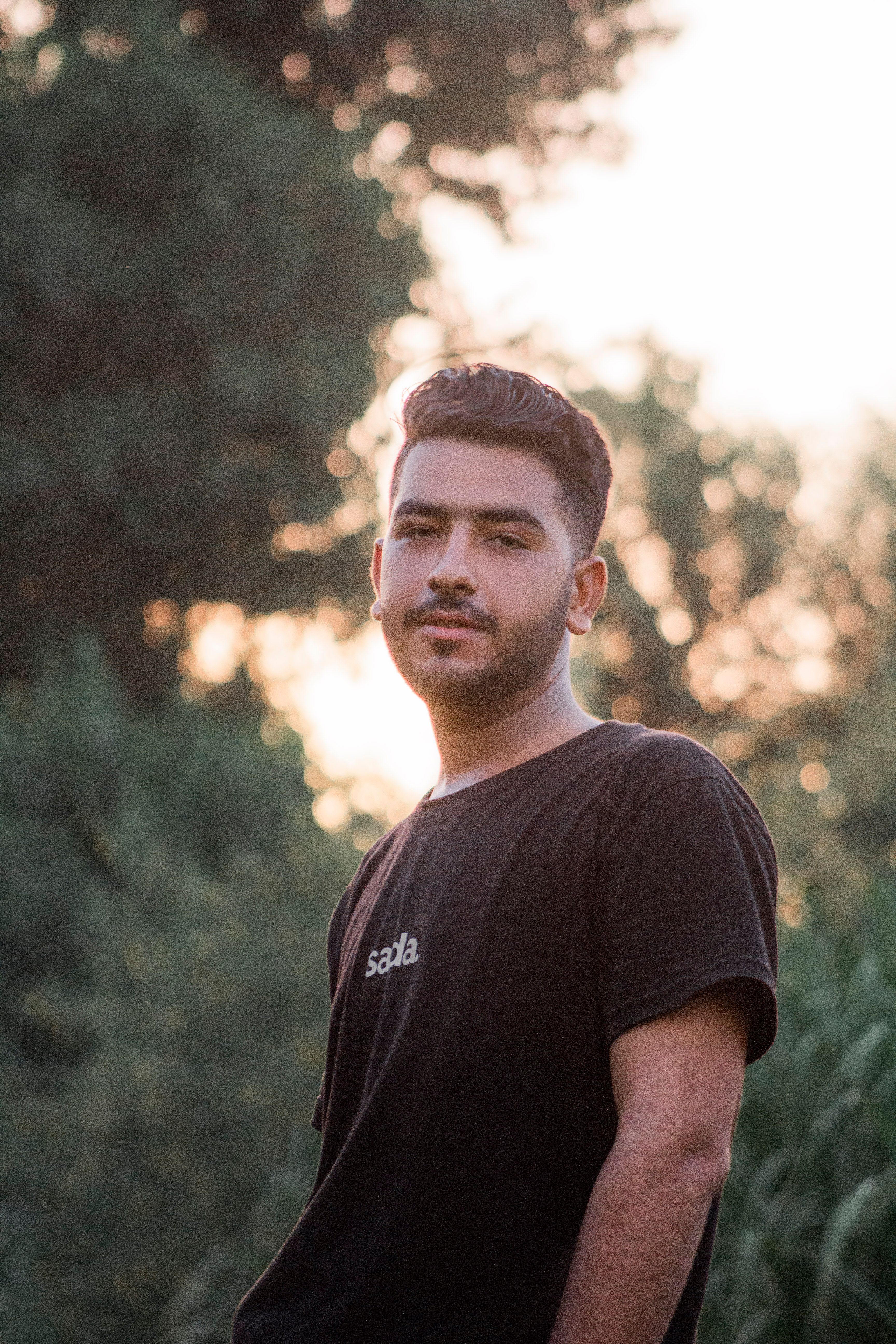 A young man stands outdoors in casual clothing, captured during a sunset with trees in the background.