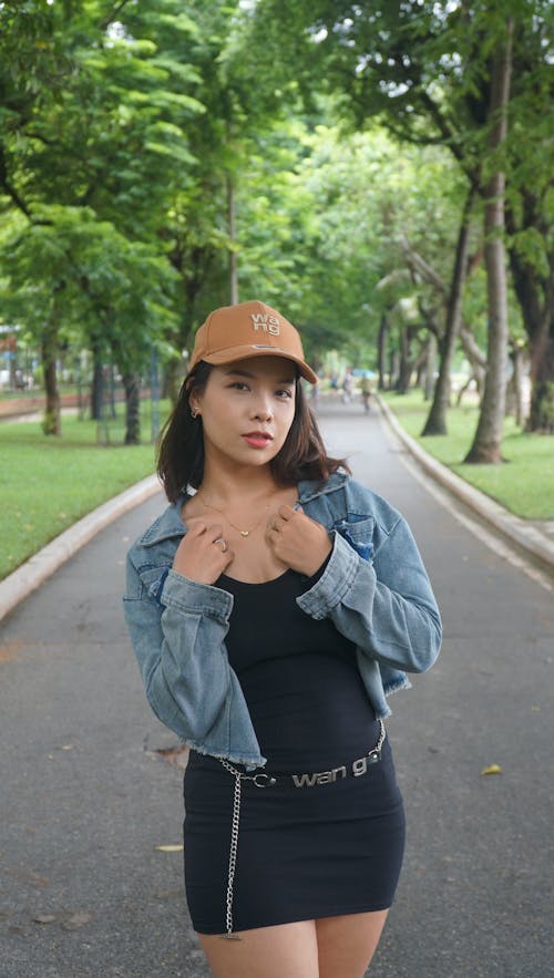 Woman in Cap and Jean Jacket Standing in Alley in Park