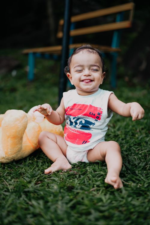 Free Adorable baby sitting on grass, laughing with a plush toy beside. Captures joy and innocence. Stock Photo