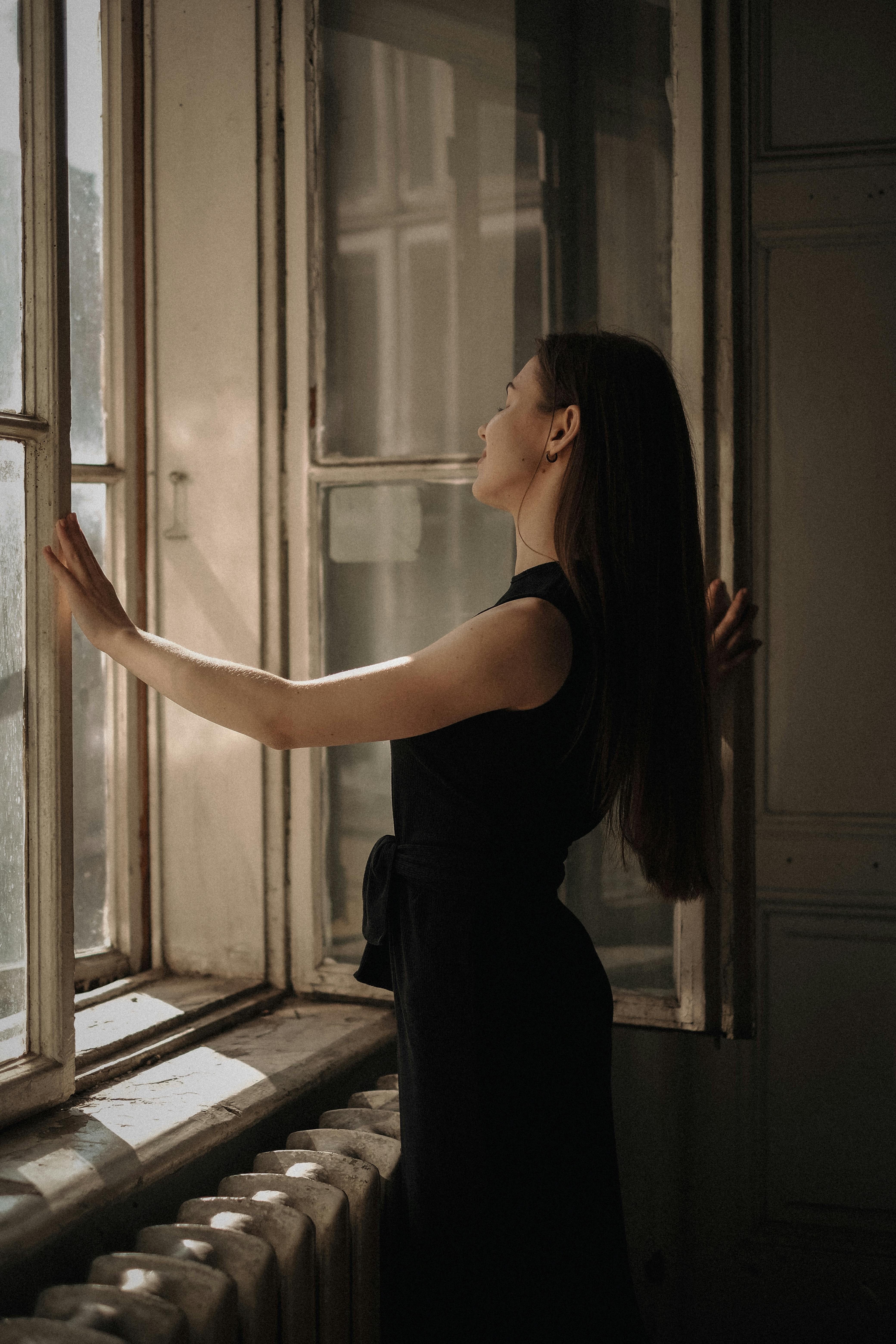 A brunette woman in a black dress stands elegantly by a sunlit window, adding sophistication to the simple room.