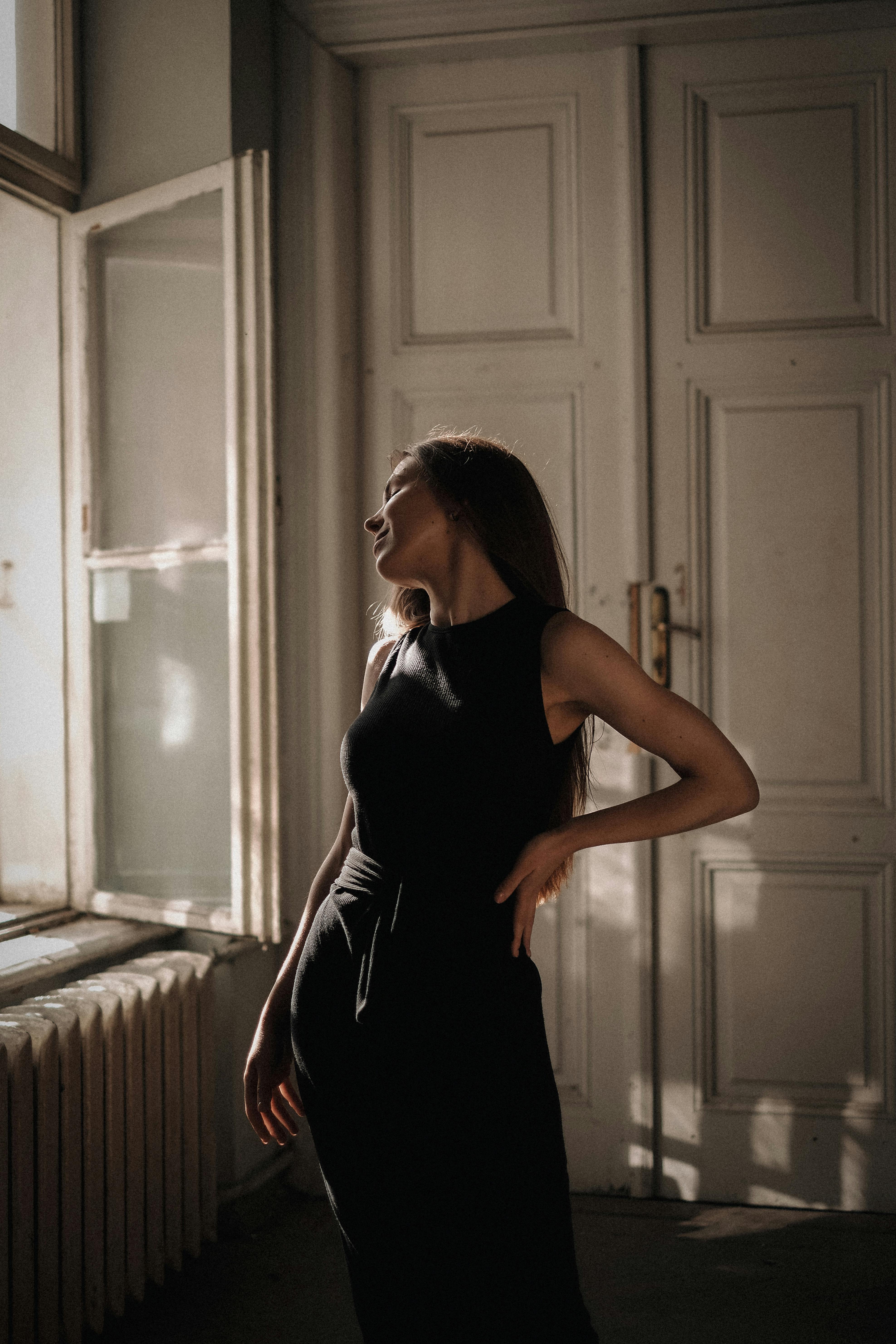 Free A woman poses gracefully in a classic room, wearing a black dress and basking in natural light. Stock Photo
