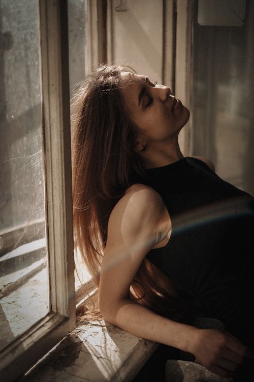 Free Serene portrait of a woman with long brown hair leaning on a sunlit windowsill. Stock Photo