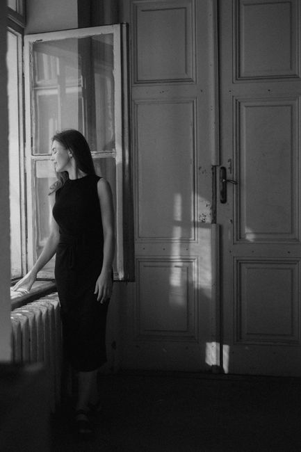 A stylish woman in a black dress stands by an open window, casting a dramatic shadow in a vintage room.