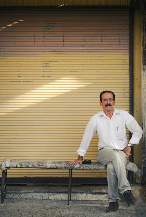 A Man with Mustache Sitting on a Bench in front of a Building 