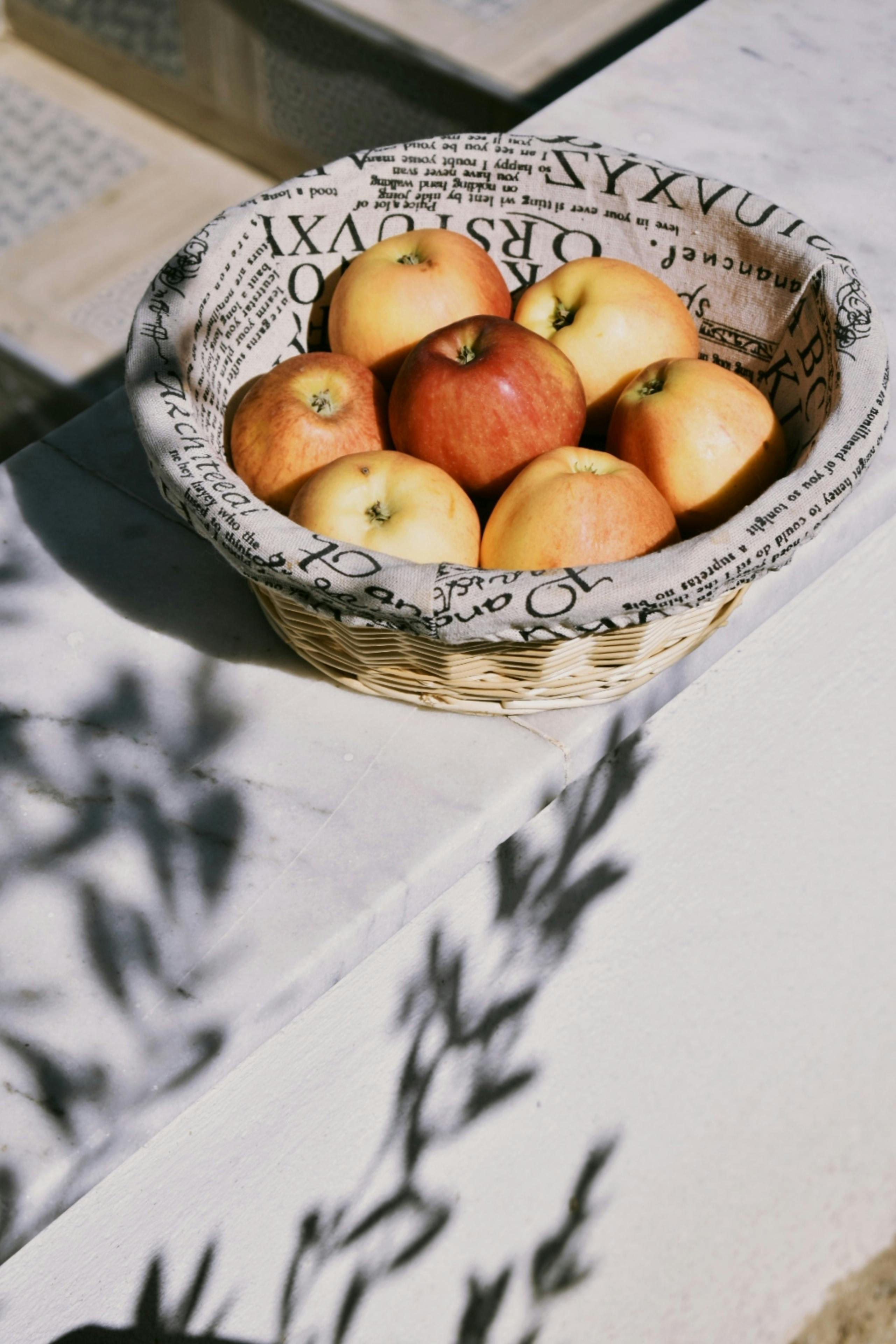 A wicker basket with fresh apples on a sunlit marble table, surrounded by newspaper lining.