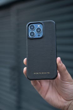 Close-up of a hand holding a smartphone in a black textured case with a blurred background.