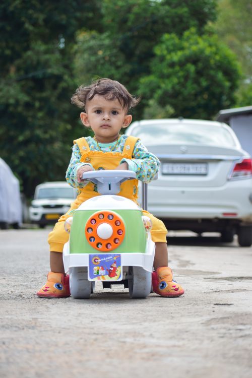 Free A young child rides a toy car on an urban street in Korba, India, surrounded by parked vehicles. Stock Photo