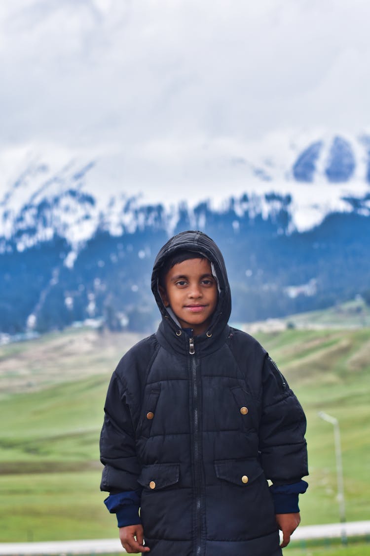 Teenager Boy In Black Hooded Puffer Jacket Posing With Snowy Mountain Peaks In Background