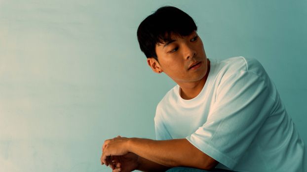 Thoughtful man looking down against a soft blue background, wearing a white t-shirt.