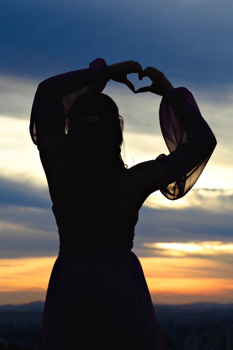 Silhouette Of Woman Standing And Showing Heart Gesture At Sunset