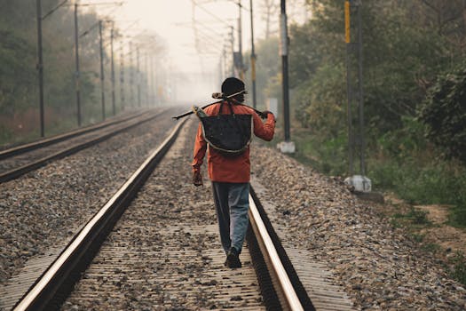 Alone traveler walking on railway tracks carrying bag on stick. Captured from behind.