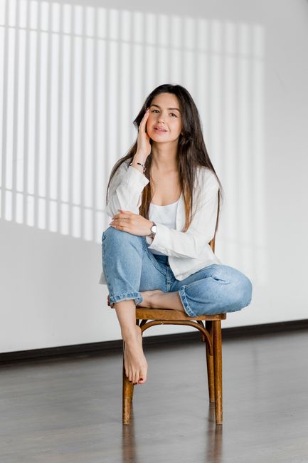 Casual portrait of a smiling woman sitting barefoot on a chair indoors with natural light.