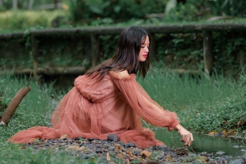 Free A woman in a flowing dress sits beside a serene stream in a lush, rustic setting. Stock Photo