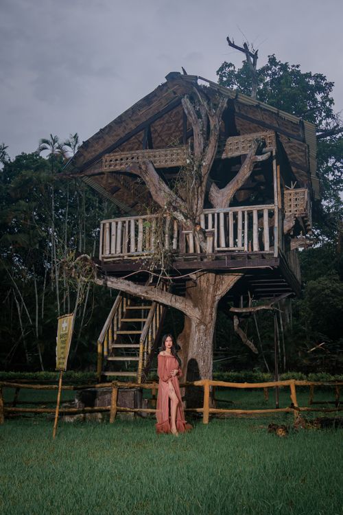 Free A serene image of a woman standing near a rustic treehouse surrounded by lush greenery. Stock Photo
