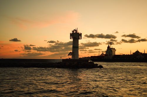 Free Silhouette of a lighthouse at sunset with a calm harbor and sky reflecting warm hues. Stock Photo