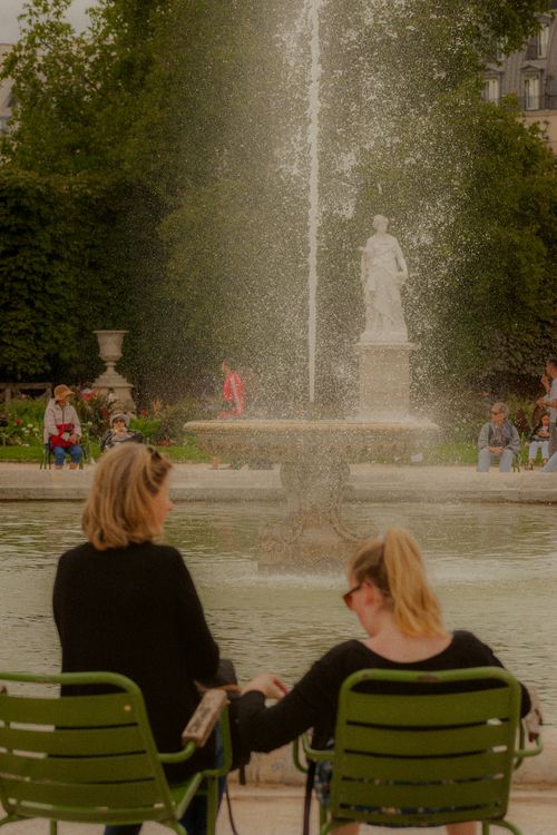 Free Two women sit by a fountain statue in an urban park, enjoying a tranquil day. Stock Photo