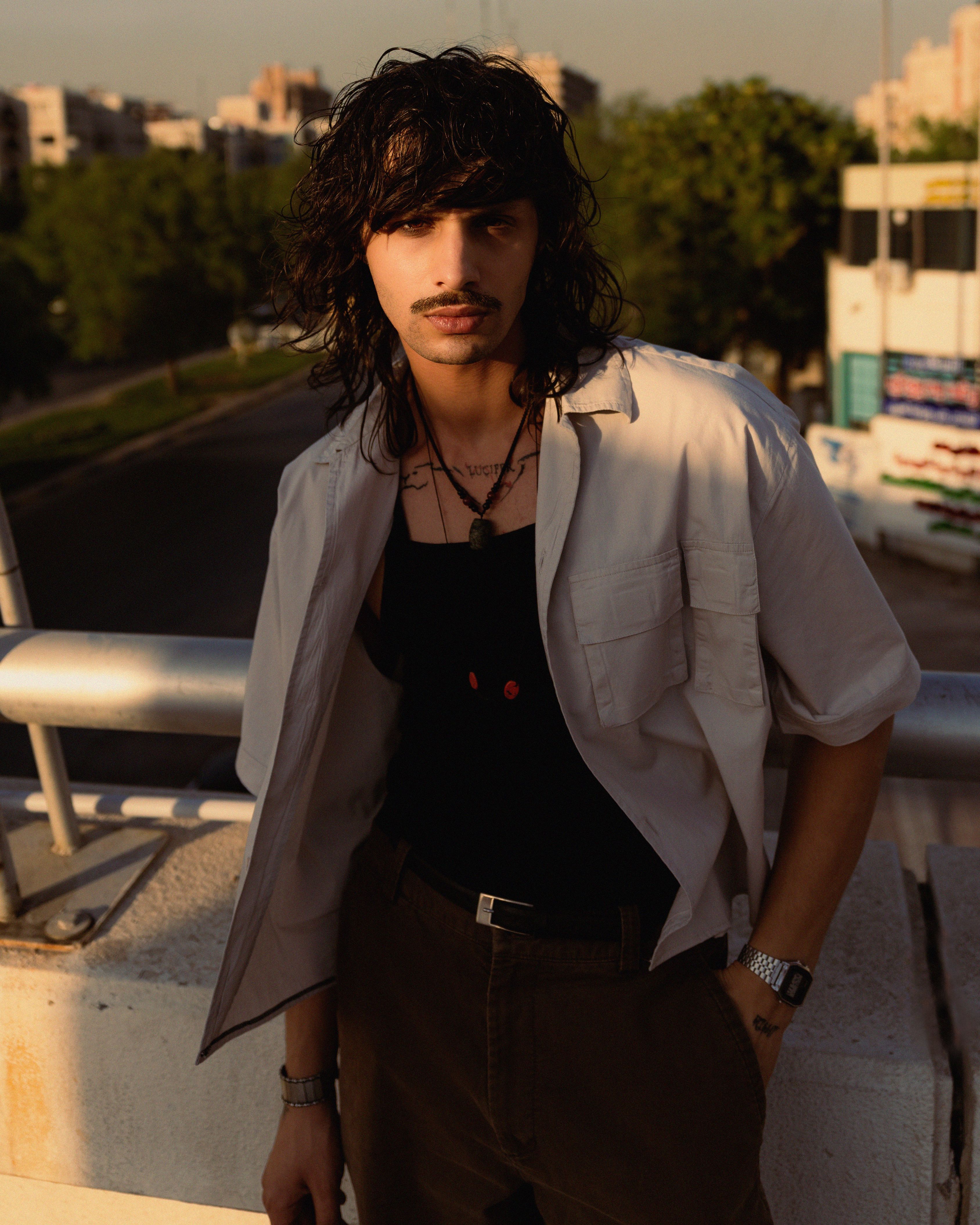 Stylish man with long hair posing outdoors in an urban setting with a relaxed stance.