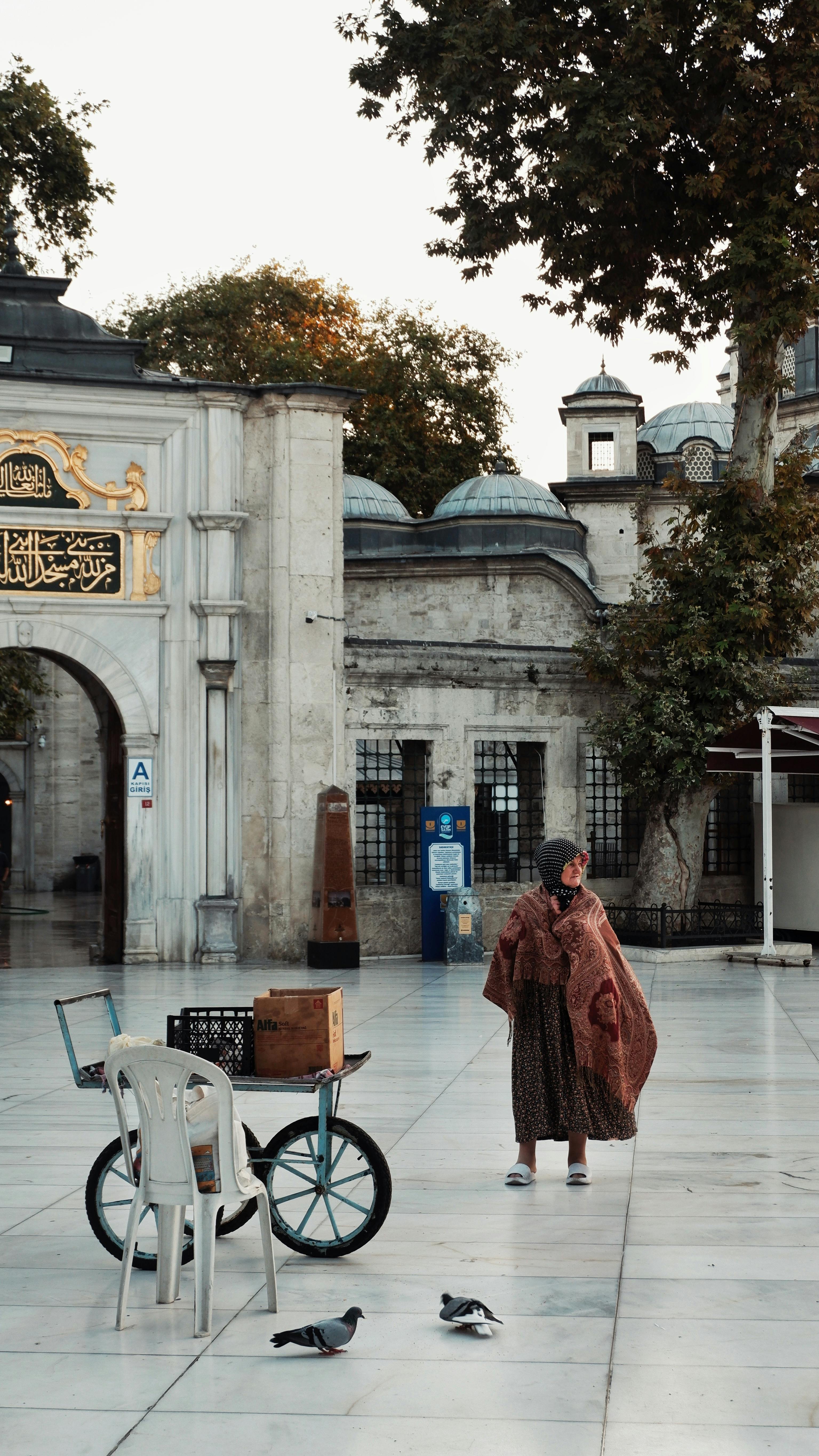 An elderly woman stands near a bicycle in a historic square, framed by Islamic architecture.