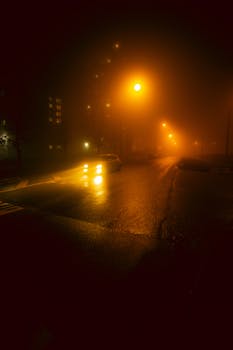Car headlights illuminate a foggy city street at night, creating a moody atmosphere.