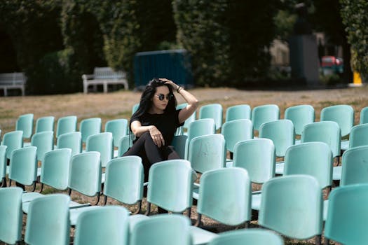 A fashionable woman in black sits among empty turquoise seats outdoors.