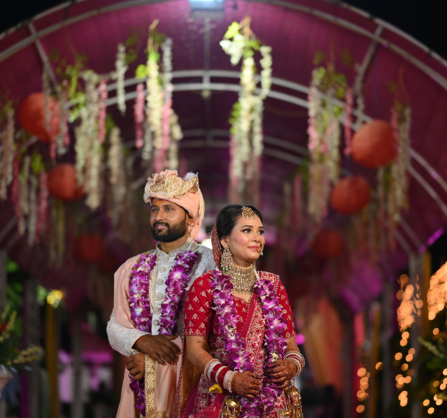 Newlyweds in traditional Indian attire standing under a decorated pavilion at night.