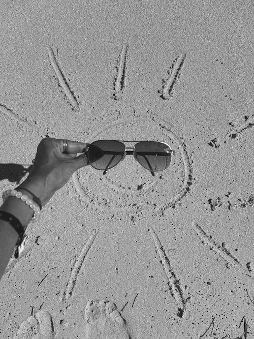 Hand Holding Sunglasses over Sun Face on Sand