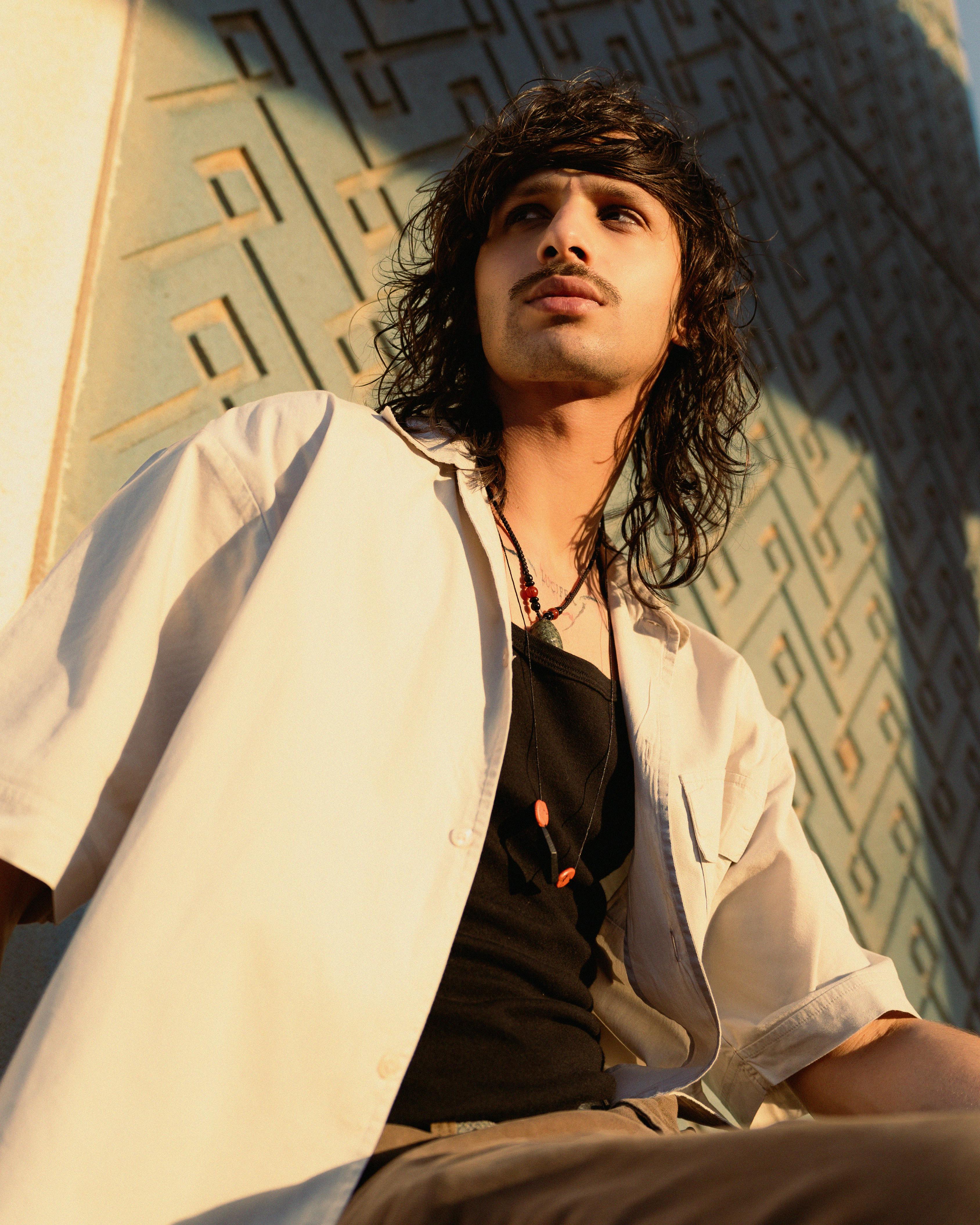 Dynamic portrait of a young man with long hair posing against an architectural wall in sunlight.