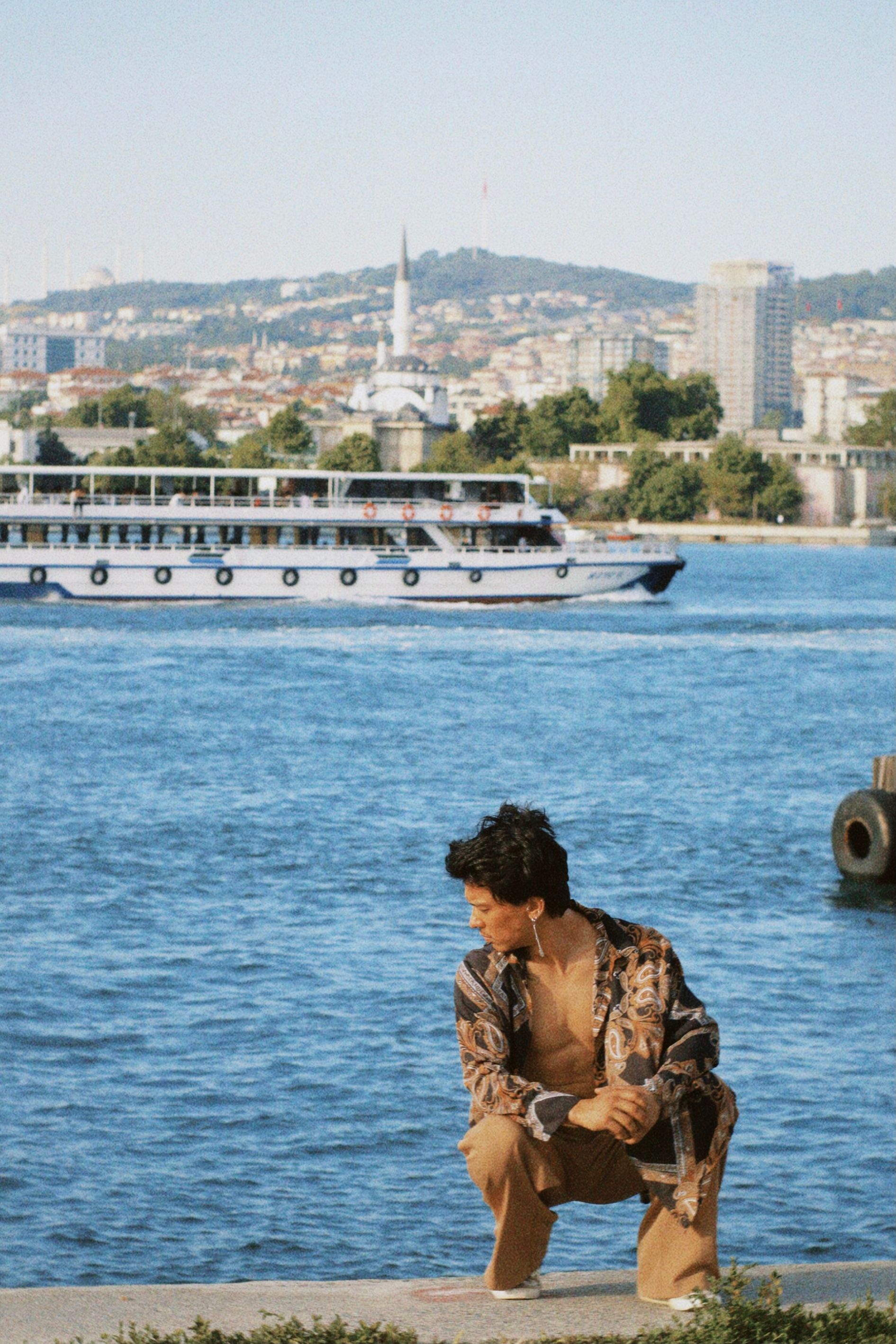 Man squatting by a riverside with a cityscape and ferry in the background. Serene urban scene.