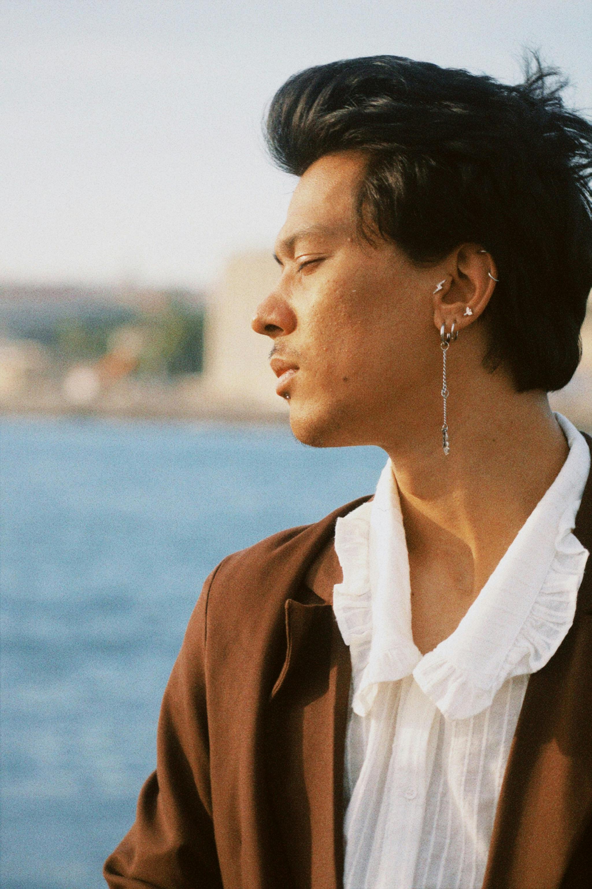 Stylish man with earrings, eyes closed, by the sea wearing a white shirt and brown blazer.