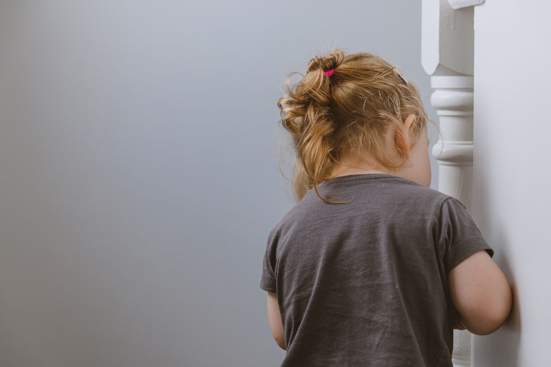 A young girl with a ponytail plays hide and seek, facing a corner inside the house.