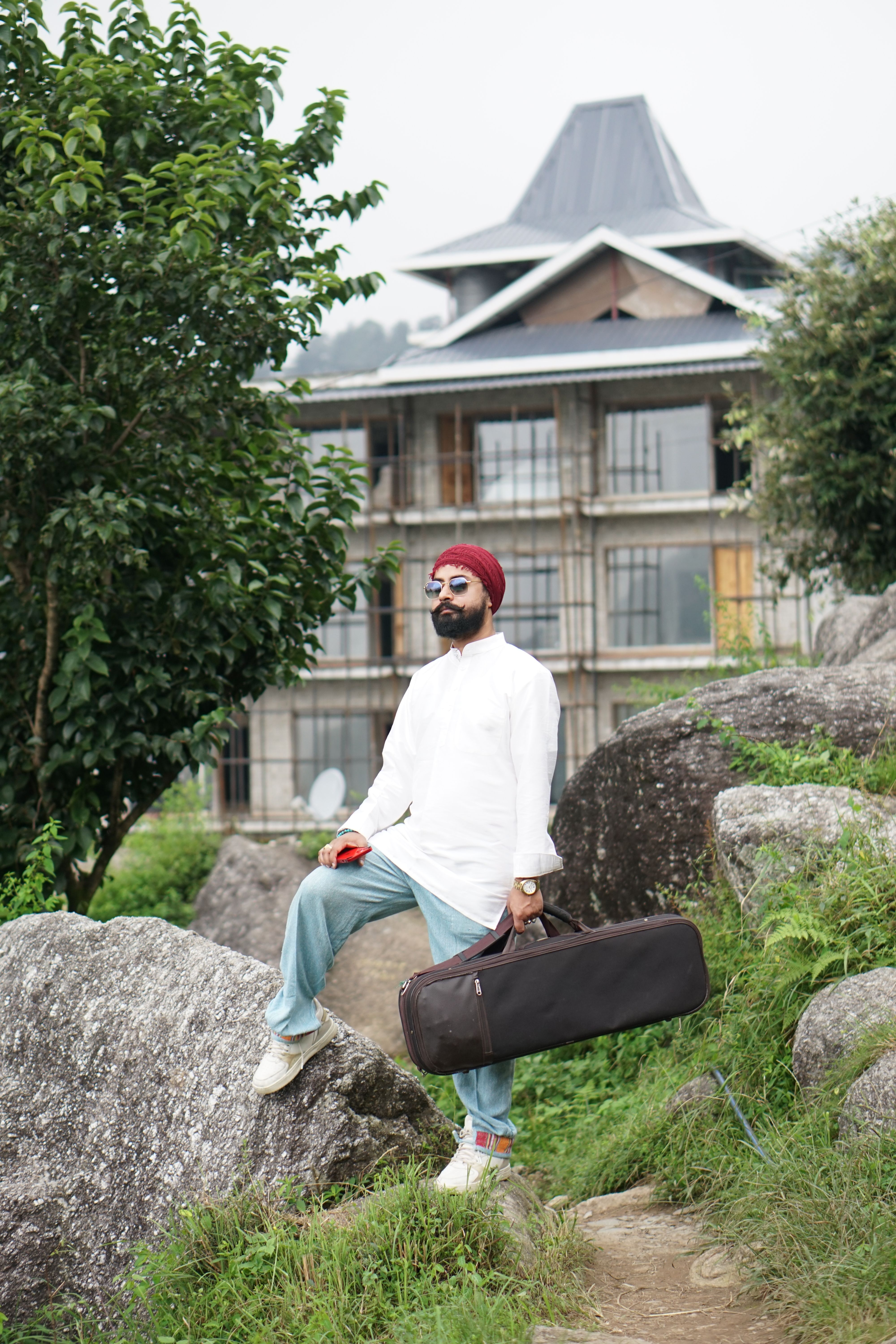 Man with a beard and turban holding a guitar case in an Indian townscape.