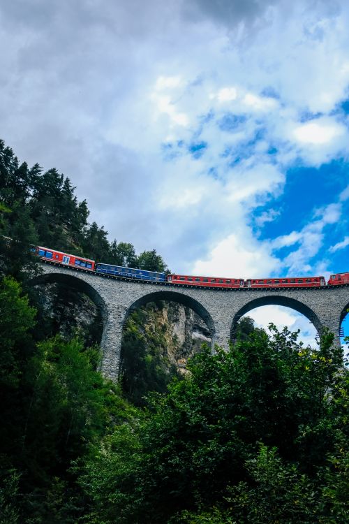 Free A vibrant train traveling over an arched bridge surrounded by lush greenery and blue sky. Stock Photo