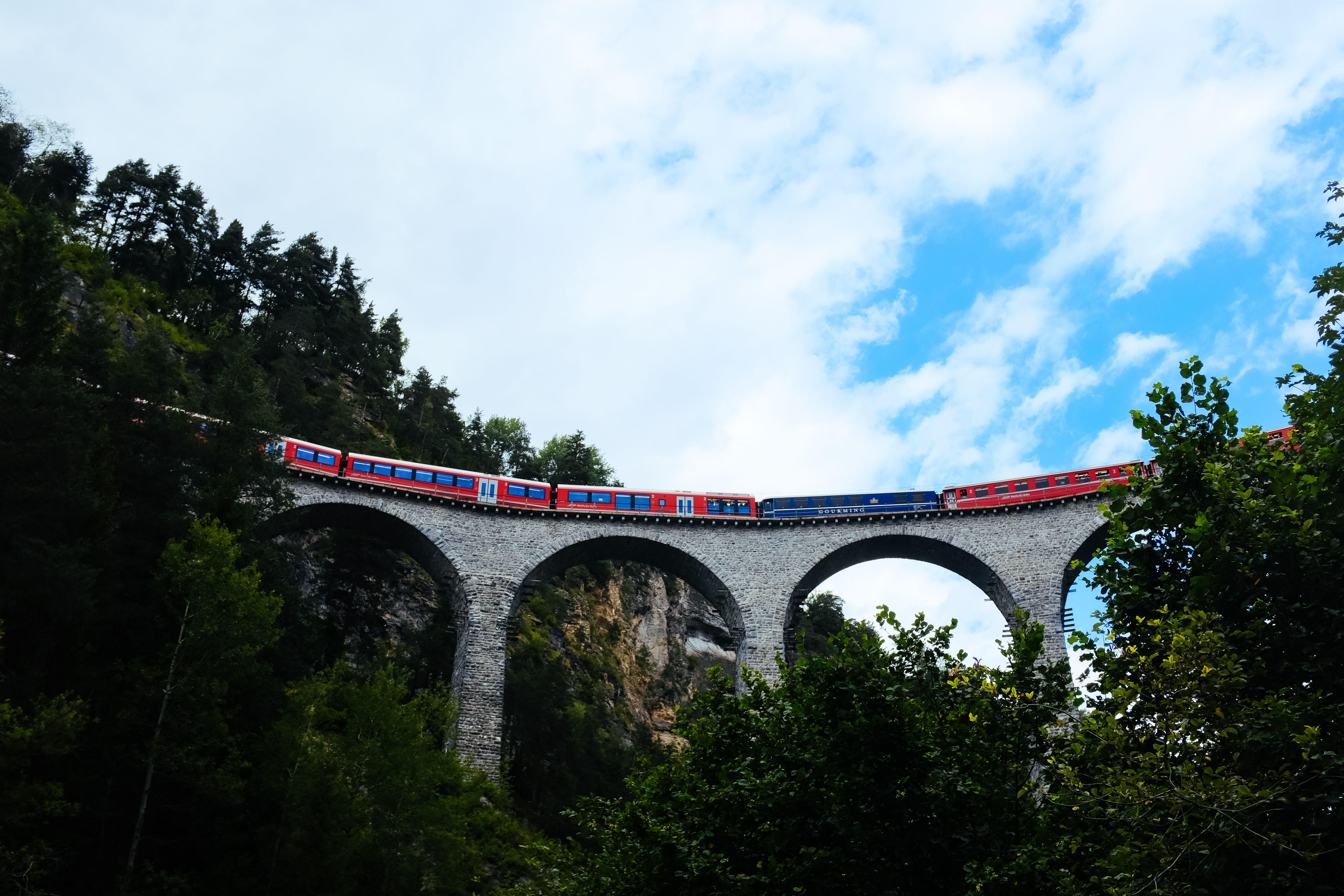 A vibrant red train crosses an arched stone bridge amidst lush greenery and blue sky.