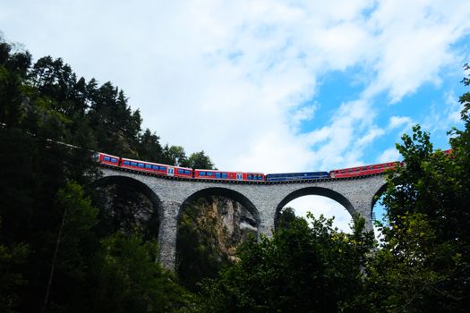 A vibrant red train crosses an arched stone bridge amidst lush greenery and blue sky.