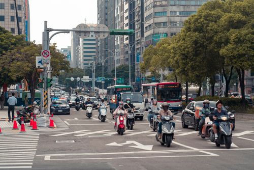 Free People riding scooters and vehicles at a busy urban intersection with traffic lights and city buildings. Stock Photo