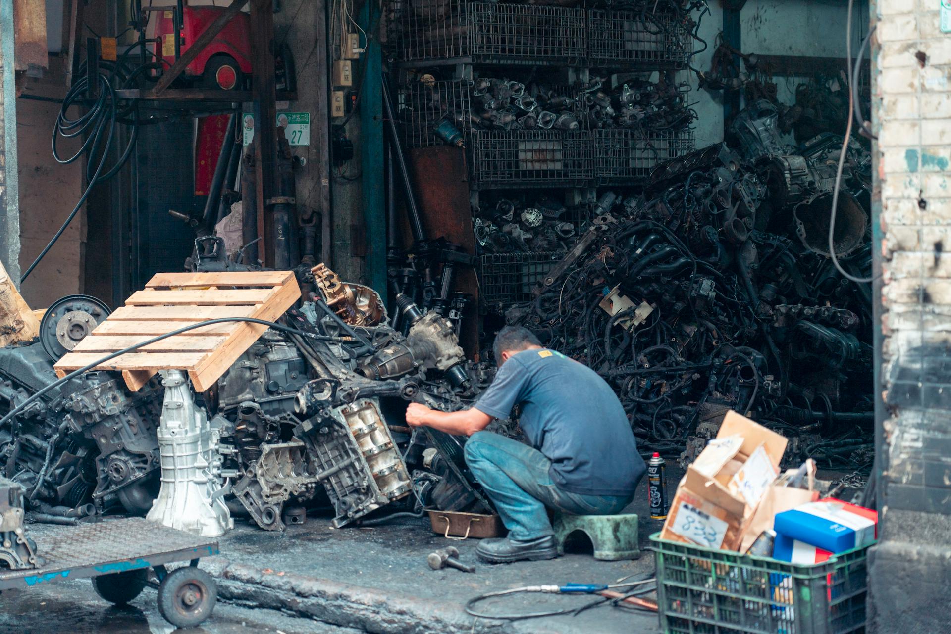 Mechanic fixing auto parts in a cluttered urban workshop, surrounded by machinery.