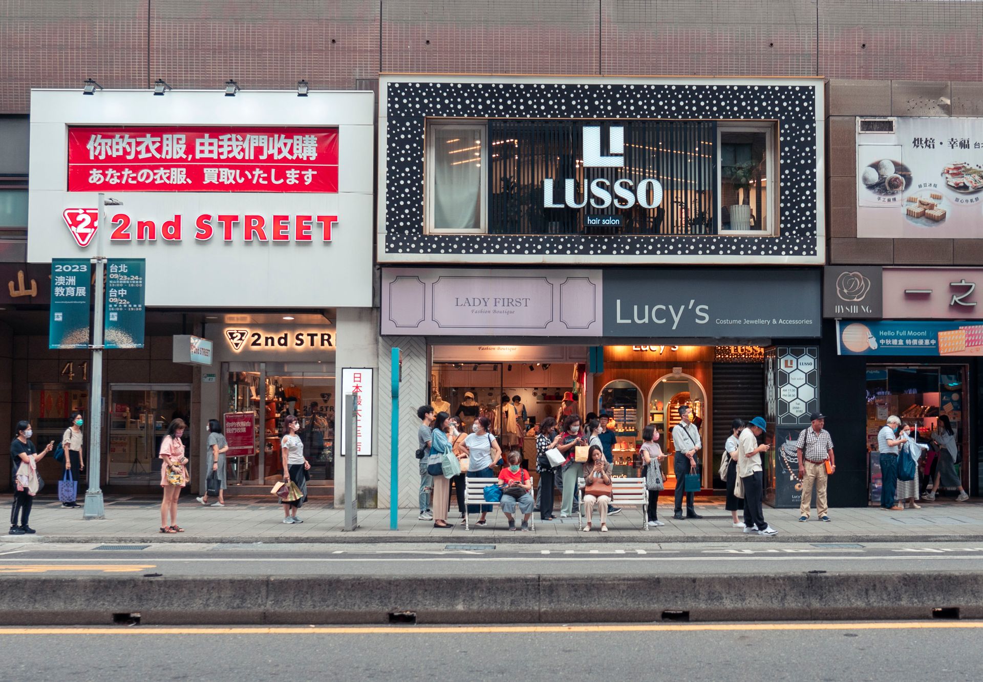People waiting at a bus stop on a busy street in Taipei, displaying urban life and local businesses.