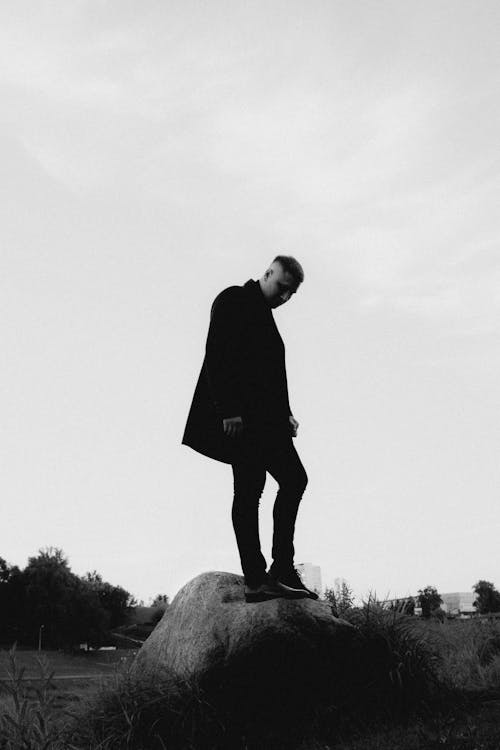 a man in a black coat, black pants and shoes stands on a rock and looks downward