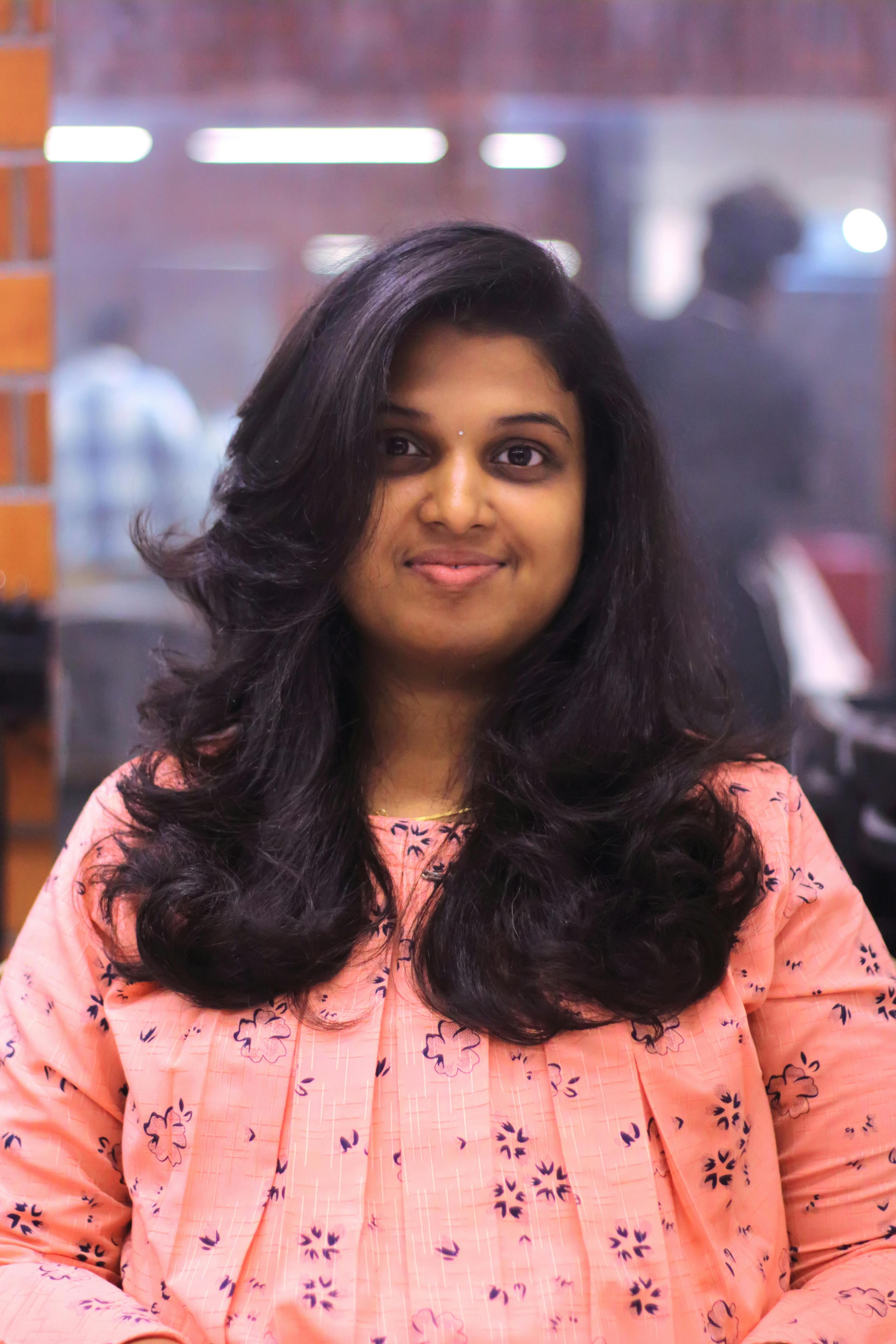 Portrait of a smiling woman with long dark hair, wearing a pink patterned top, sitting indoors.