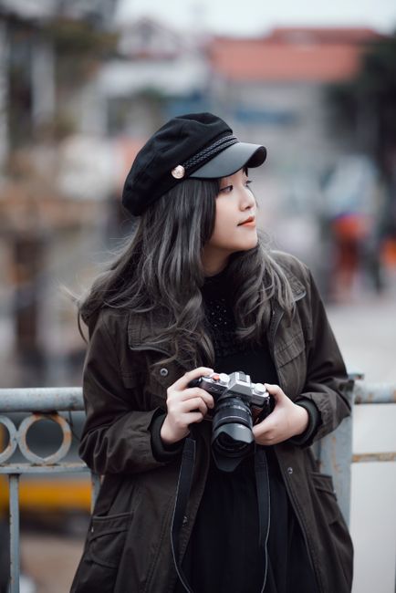 Asian woman in fashionable attire with camera, standing outdoors in urban setting.