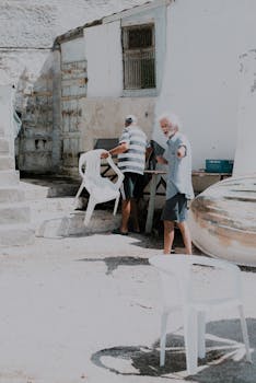Two adult men arranging white plastic chairs outside in a sunny alley.