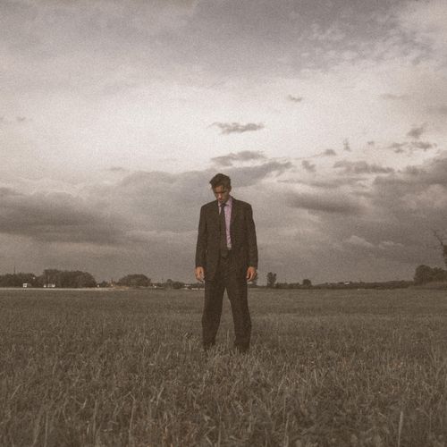 Free A man in a suit standing alone in a vast grassy field under a stormy sky. Stock Photo