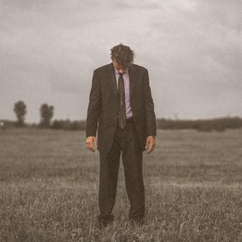 Free A man in a suit reflects in solitude, standing in a grassy field under a cloudy sky. Stock Photo