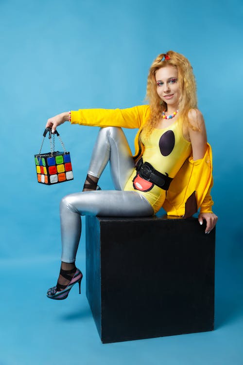 Woman Wearing Yellow Jacket And Grey Pants Sitting On Black Cube